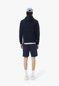 Felpa blu navy con cappuccio regolabile e tasca a marsupio, abbinata a pantaloni corti coordinati, t-shirt bianca, calze bianche e sneakers bianche.