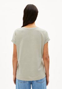 Camicetta a maniche corte, di un verde chiaro, realizzata in morbido cotone, con un taglio rilassato e risvolti sui polsini. Vista posteriore, abbinata a jeans di denim blu.