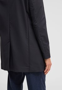 Mann trägt einen schwarzen Mantel mit langen Ärmeln und Rücken-Schlitz, steht mit einer Hand entspannt an seiner Seite und trägt dunkelblaue Hosen.