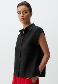 Camicia nera di lino con colletto, maniche corte, apertura con bottoni e spacchi laterali. Indossata con pantaloni rossi, il tessuto ha una finitura testurizzata.