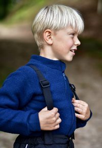 Junge mit kurzem, hellblondem Haar trägt eine blaue Fleecejacke mit Reißverschluss und schwarze Träger, die dunkle Hosen halten. Natürlicher Hintergrund.