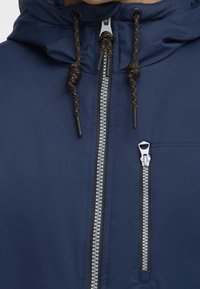 Giacca blu navy con cappuccio, caratterizzata da una cerniera argento e una tasca sul petto con zip. Stringhe testurizzate con accenti scuri e chiari.