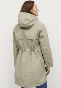 Lichtbeige waterdichte jas met een capuchon, taille met trekkoord en een ventilerende rughals. Heeft een subtiel logo op de capuchon. Lange mouwen.
