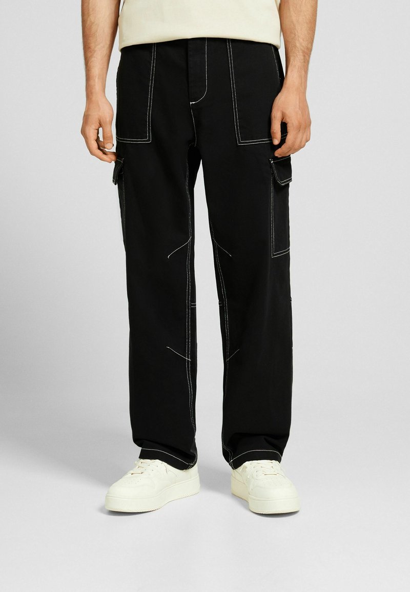 Pantalon cargo noir avec coutures blanches, doté de poches latérales et d'une coupe ample. Associé à des baskets blanches et un haut de couleur claire.