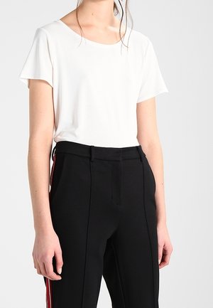 Frau trägt eine weiße kurzärmlige Bluse, die in schwarze, hoch taillierte Hosen mit roten und weißen Seitenstreifen gesteckt ist.