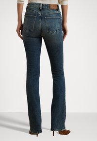 Temno modri flare jeans z visokim pasom, obogateni s svetlo rjavim usnjenim logotipom na hrbtu in klasičnim oblikovanjem žepov.