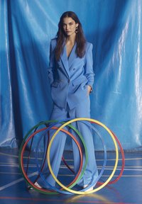 Light blue tailored suit with double-breasted jacket and wide-leg trousers, paired with white shoes. Colorful hula hoops arranged in front.