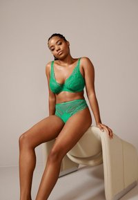 Reggiseno verde in pizzo e coordinato slip alto con dettagli in rete. Tessuto dalla consistenza liscia e motivi floreali intricati. Modella seduta su una sedia color crema.