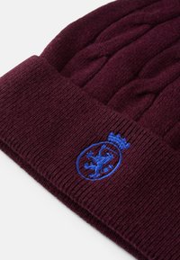 Burgundowa dziana czapka z teksturowanym ściągaczem i niebieskim haftowanym emblematem z motywem lwa i korony.