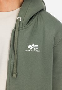 Χακί φούτερ με φερμουάρ, κατασκευασμένο από μίγμα βαμβακιού, με κουκούλα ρυθμιζόμενη με κορδόνι και λευκό λογότυπο "Alpha Industries" στο στήθος.