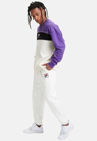Pantaloni tuta bianchi con polsini elasticizzati, con logo FILA; abbinati a un pullover girocollo a blocchi di colore viola e bianco.