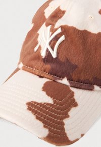 Căciulă de camuflaj maro și bej, realizată din țesătură moale, având un logo alb brodat pe față și o vizieră curbă cu detalii cusute.