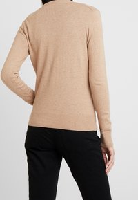 Beige stickad tröja med ribbad nederkant och ärmslut, långa ärmar och hög hals, kombinerad med svarta byxor, sedd bakifrån.