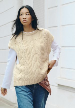Donna con i capelli lunghi e neri che indossa un maglione smanicato lavorato a maglia color crema sopra una maglietta bianca a maniche lunghe e jeans blu, mentre cammina all'aperto.