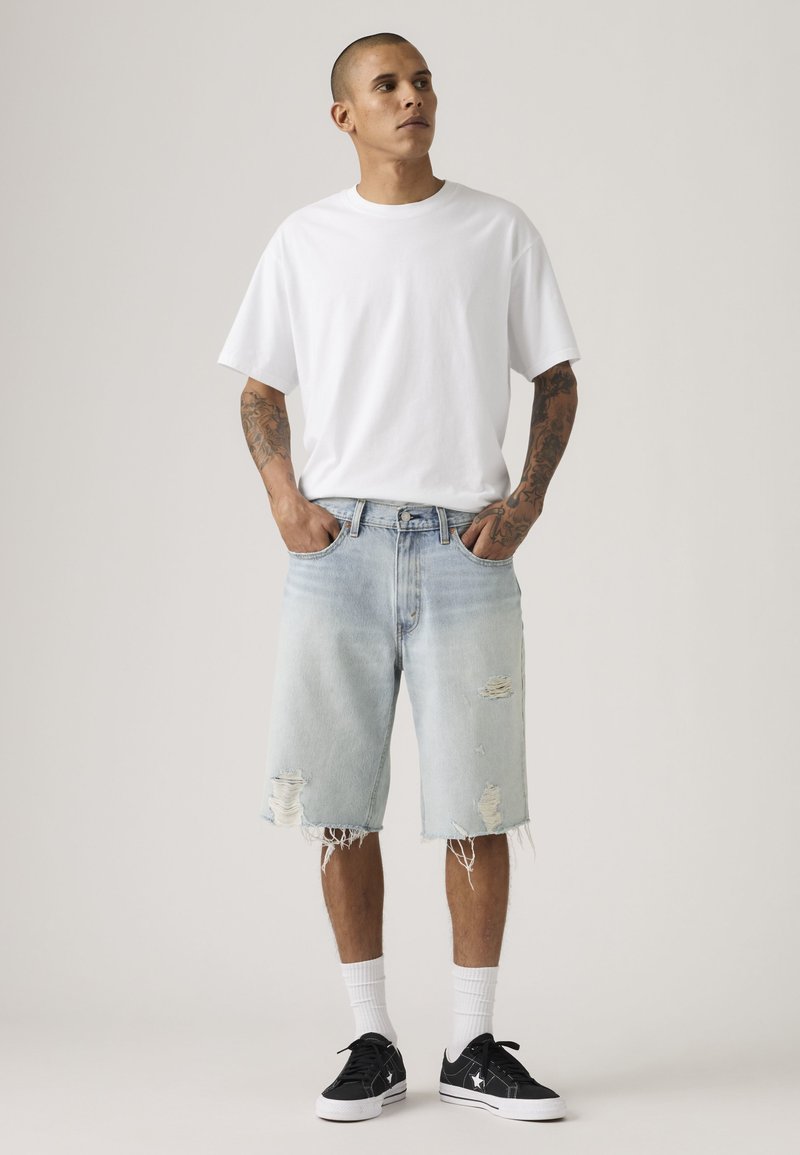 Man met tatoeages draagt een wit oversized T-shirt, licht versleten denim shorts, witte sokken en zwarte sneakers, staand met handen in zijn zakken.