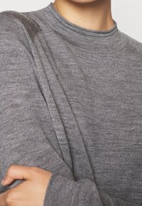 Person, der einen grauen, gestrickten Pullover mit langen Ärmeln trägt, mit einem Arm über die Brust verschränkt und die Hand am gegenüberliegenden Ärmel haltend.