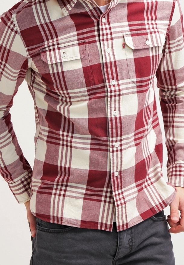 Homme portant une chemise à manches longues à carreaux rouges et blancs avec deux poches poitrine et un pantalon noir.