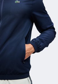 Άνδρας με ναυτικό μπλε μπουφάν Lacoste με το χέρι στην πλαϊνή τσέπη και ελαστικά μανίκια ορατά στα μανίκια.
