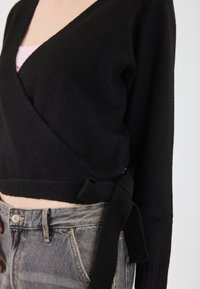 Czarny sweter opasany wykonany z miękkiego materiału, z głębokim dekoltem w serek, długimi rękawami i wiązaniem z boku. Połączony z szarymi dżinsami o wysokim stanie.