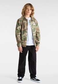 Tarnjacke in Grün-, Braun- und Beige-Tönen, ein weißes T-Shirt darunter, schwarze Hose und schwarze Vans-Schuhe.