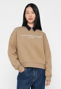 Beige genser med blå krage, som har hvit "TOMMY HILFIGER EST. 1985" logo. Løs passform med ribbede mansjetter og hem, kombinert med blå jeans.