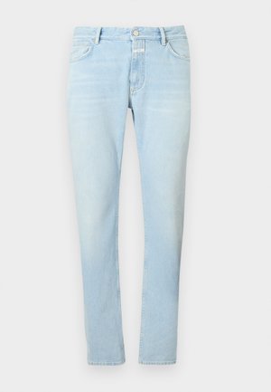 Junger Mann, der ein schwarzes langärmeliges Hemd, hellblaue Jeans, schwarz-weiße Loafers und eine beige Umhängetasche trägt, steht mit einer Hand in der Tasche.