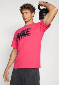 Rosa Nike T-shirt med sort logo og tekst, med korte ærmer og rund hals. Bærende med mørke teksturerede shorts, holder en kettlebell.