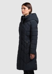 Vrouw die een lange zwarte gewatteerde jas draagt met een grote capuchon, staand in profiel tegen een effen lichte achtergrond.