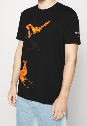 Mann trägt ein schwarzes T-Shirt mit einem großen orange-schwarzen Cartoon-Enten-Motiv auf der Vorderseite und hellblaue Jeans.
