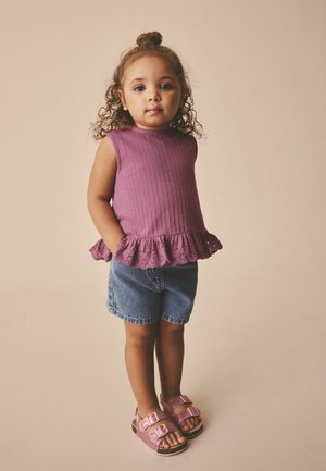 Young child with curly hair in a purple sleeveless top, denim shorts, and pink sandals standing against a plain beige background.