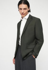 Dunkelgrüner Blazer aus strukturiertem Stoff, mit einer einzigen Brusttasche, zwei Fronttaschen und silbernen Knöpfen. Kombiniert mit einem weißen Hemd.