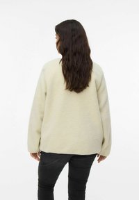 Chaqueta de forro polar color crema con forma cuadrada, mangas largas y una textura suave. Lleva puesta sobre pantalones negros, mostrada desde la parte trasera.