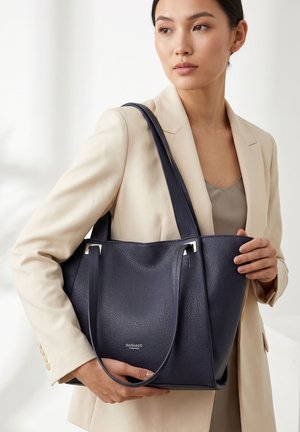 Femme en blazer beige tenant un grand sac cabas en cuir bleu marine avec des ferrures argentées et des doubles bretelles.
