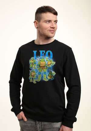 Uomo con felpa nera con tartarughe dei cartoni animati che tengono armi e testo "LEO", in piedi contro uno sfondo semplice, guarda verso destra.