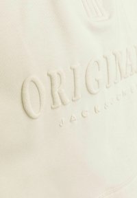 Krém színű szövet, texturált felülettel, kiemelkedő, hímzett szöveggel, ami így szól: "ORIGINAL JACK & JONES." Egyszerű, diszkrét dizájn.
