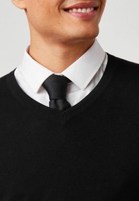 Suéter negro de cuello en V, hecho de un material de punto suave, llevado sobre una camisa blanca con cuello y una corbata negra de canalé. Vista en primer plano del área del cuello.