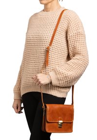 Borsa a tracolla in pelle marrone con pattina e hardware dorato, abbinata a un maglione lavorato a maglia beige e pantaloni neri.