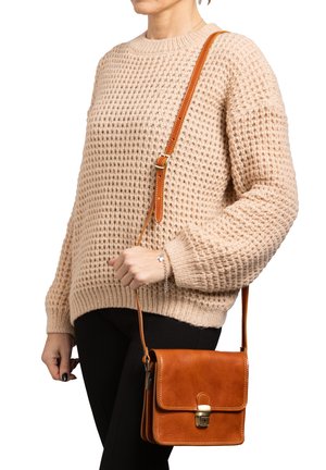 Borsa a tracolla in pelle marrone con pattina e hardware dorato, abbinata a un maglione lavorato a maglia beige e pantaloni neri.