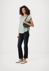 Sieviete ar lokainiem matiem, ģērbta piparmētru zaļā augšdaļā, tumšās džinsās, bezdelīgu bežās kurpēs un turot nelielu brūnu pleca somu, stāv uz balta fona.