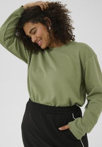 Sweatshirt vert olive avec un col rond et des manches longues, associé à un pantalon de survêtement noir doté d'une taille élastique et d'accents sur les côtés.