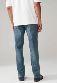 Jeans de mezclilla azul con un diseño de corte recto, que cuentan con bolsillos y una ligera textura de lavado, combinados con zapatos a rayas marrones y azules.