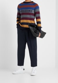 Gestreepte gebreide trui in blauw, rood en geel met "MISSONI" logo. Gecombineerd met marineblauwe pantalon met krijtstrepen en witte sneakers, en een zwarte tas in de hand.