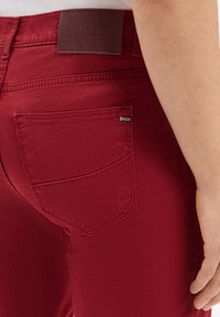 Gros plan sur un pantalon Brax rouge montrant la poche arrière, une étiquette en cuir de la marque, et une partie de la main d'une personne reposant à côté de la hanche.