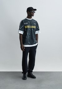 Άνδρας με μαύρο καπέλο, γυαλιά ηλίου, μαύρο και άσπρο πουκάμισο Moschino "Signed Garment", μαύρο παντελόνι και μαύρα αθλητικά παπούτσια στέκεται σε γκρι δάπεδο.