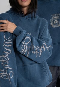 Hoodie en coton bleu avec une capuche à cordon. Présente un texte brodé gris sur les manches et un petit logo sur la poitrine. Texture douce.