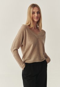 Pull beige à col en V avec poignets côtelés et coupe ample, fabriqué en tissu doux. Associé à un pantalon noir ample.