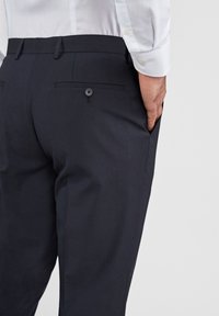 Marineblauwe broek met een zachte textuur, voorzien van twee achterzakken met knoopsluiting en een slim fit ontwerp. Combinatie met een lichtgekleurd shirt.
