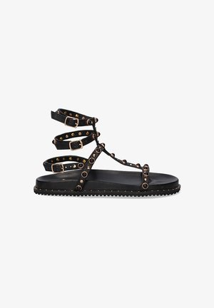 Sandalias planas negras con múltiples correas ajustables, que presentan tachuelas doradas a lo largo de las correas y una suela texturizada. Diseño minimalista y material duradero.