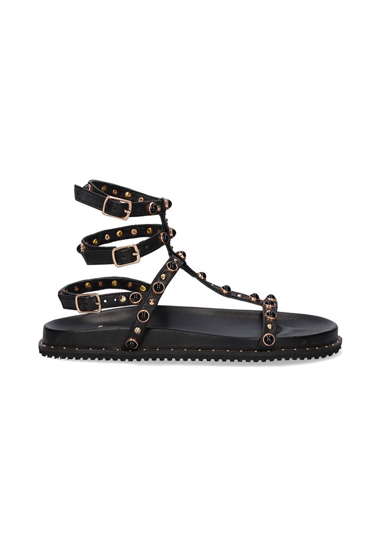 Sandalias planas negras con múltiples correas ajustables, que presentan tachuelas doradas a lo largo de las correas y una suela texturizada. Diseño minimalista y material duradero.