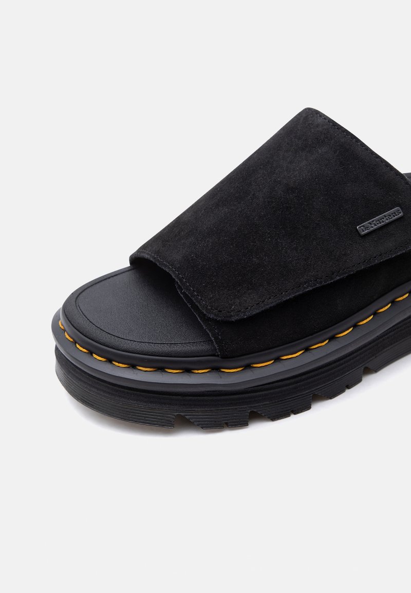 Sandalo a ciabatta in suede nero con suola piatta spessa, cuciture gialle intorno alla suola intermedia e una piccola targhetta metallica con il marchio sulla fascia superiore.